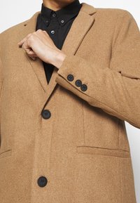Blazer marrone in misto lana con rever a punta, tre bottoni neri e tasche laterali, indossato su una camicia nera. Texture liscia.