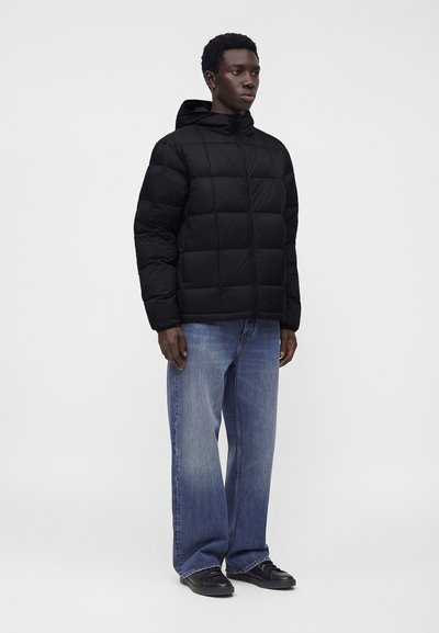 Jeune homme portant une doudoune noire à capuche, un jean bleu ample et des chaussures noires, debout devant un fond clair uni.