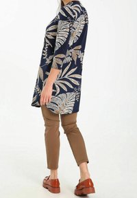 Granatowa sukienka tunika z białym i złotym wzorem kwiatowym, z rękawami o długości trzy czwarte, połączona z brązowymi dopasowanymi spodniami i brązowymi butami na koturnie.