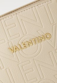 Beige textúrájú bőrfelület, amelyen arany színű "VALENTINO" logó van debütálva, feltűnő betűstílussal és diszkrét mintákkal.