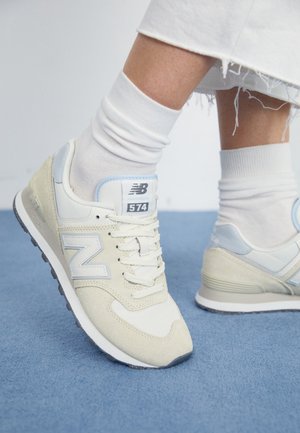 Ben iført hvide sokker og lyse beige New Balance 574 sneakers stående på blåt tæppe med flossede hvide bukser.