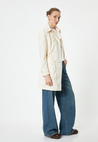 Trench coat doppiopetto color crema chiaro con vita regolabile, abbinato a jeans in denim blu a gamba larga e scarpe chunky marroni.