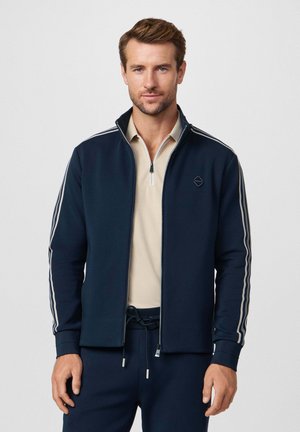Chaqueta azul marino con cremallera y rayas blancas contrastantes en las mangas. Tejido suave, cuello alto y un logo en forma de diamante en el pecho. Lleva una camiseta beige a juego debajo.
