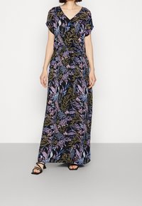 Femme portant une robe maxi noire avec des motifs floraux violets, bleus et olive, des manches courtes, un décolleté en V et des sandales noires à lanières.