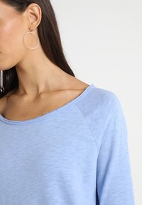 Vaaleansininen palloaukkoesine pehmeällä, teksturoidulla kankaalla ja raglanhihnoilla. Yhdessä, hohtava renkaankorvisten kanssa.