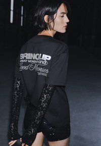 Črna majica s kratkimi rokavi z belim besedilom na hrbtnem delu, združena z dolgimi črnimi čipkastimi rokavi in krilom iz bleščic. Temno ozadje z mehko osvetlitvijo.