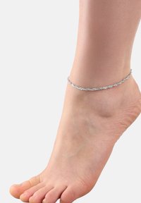 Tobillera de plata con delicados eslabones de cadena entrelazados, llevada en un pie descalzo, resaltando su diseño elegante y textura metálica.