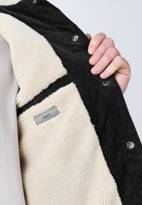 Chaqueta de pana negra con forro de lana crema, cuenta con botones a presión y una etiqueta que dice "NEXT" en el bolsillo interior.