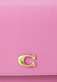 Pink leather wallet featuring a gold letter "C" emblem, smooth texture, and neat stitching along the edges. Compact rectangular design.