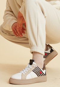 Sneaker mit einem weißen synthetischen Obermaterial, braunen Akzenten, schwarzen Streifen und einem roten Herzdetails. Gummisohle und Schnürdesign.