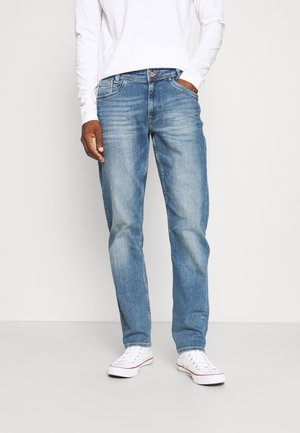 Uomo che indossa jeans azzurri sbiaditi, scarpe da ginnastica bianche e una camicia bianca a maniche lunghe, in piedi con una mano in tasca contro uno sfondo uniforme.