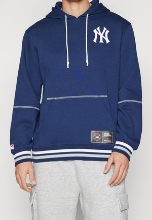 Felpa navy con logo bianco dei New York Yankees. Presenta cappuccio con cordino, polsini e orlo a righe, e un'etichetta cucita in vita. Materiale misto cotone.