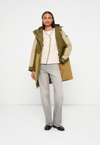 Zielona i brązowa wodoodporna kurtka z kapturem, w połączeniu z kremowym swetrem, szarymi spodniami i czarnymi butami. Posiada zatrzaski i kieszenie.