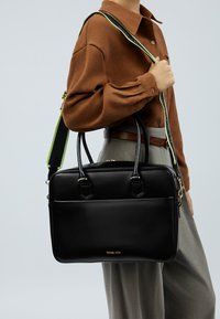 Schwarze Lederhandtasche mit zwei oberen Griffen und einem verstellbaren grünen Riemen. Verfügt über goldene Beschläge und eine glatte Textur sowie eine Fronttasche.