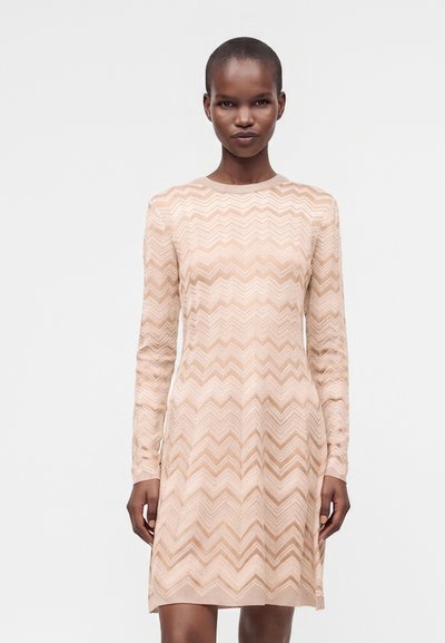Femme portant une robe beige à manches longues avec un motif en zigzag, debout devant un fond clair uni.