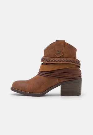 Botte cowboy marron à la cheville avec talon empilé et brides décoratives tressées et enroulées sur un fond clair.