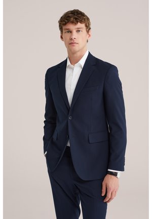 Joven con traje azul marino, camisa blanca y reloj negro, posando contra un fondo claro y sencillo.