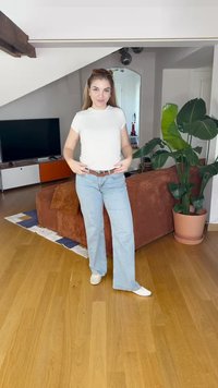 Fehér rövid ujjú póló, világoskék trapéznadrág, barna öv és fehér cipő; egy nappaliban áll, ahol egy növény és egy kanapé található.