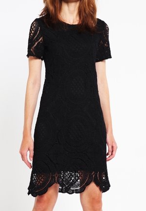 Femme portant une robe en dentelle noire à manches courtes avec des motifs circulaires et un ourlet festonné, debout devant un fond uni.