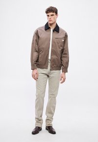 Brūna rāvējslēdzēja jaka ar melnu apkakli, divām sānu kabatām un mazu krūšu kabatu. Valkāta pāri baltai kreklam un gaiši bēšām džinsu biksēm.