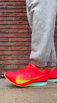 Lyserøde Nike løbesko med gule accenter, mesh-overdel og en gennemsigtig grøn sål. Grå joggingbukser er synlige ved siden af.