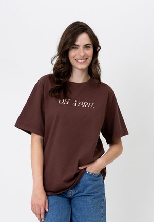 Glunderende vrouw met golvend bruin haar, gekleed in een bruin "OH APRIL" T-shirt en blauwe jeans, staand met één hand in haar zak.