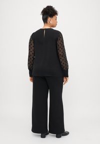 Zwarte top met doorschijnende mouwen met stippen, achterkant met sleutelgatdetail, gecombineerd met zwarte wijde broek en zwarte schoenen. Gladde stofstructuur.