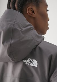 Szara kurtka z kapturem wykonana z lekkiego materiału, z wyraźnym białym logo "The North Face" na plecach, w pobliżu ramienia.