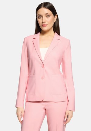 Femme portant un blazer rose clair sur mesure avec deux poches avant et un pantalon assorti, debout devant un fond blanc uni.