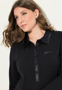 Camisa negra de manga larga con cuello de satén, frontal abotonado y un bolsillo sutil. Presenta un tejido texturizado y una silueta ajustada.