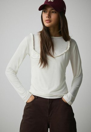 Jonge vrouw met lang donker haar, een bordeauxrode pet, een witte gelaagde top met lange mouwen en donkerbruine broek, handen in de zakken, kijkt opzij.