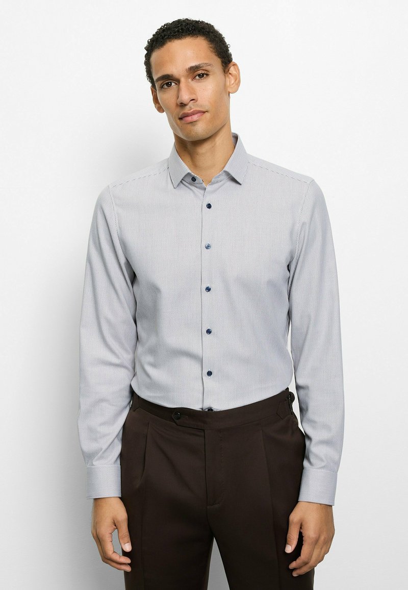 Hellblaues Langarmhemd mit feinem Streifenmuster, Button-Down-Kragen und marineblauen Knöpfen, kombiniert mit dunkelbrauner Hose.