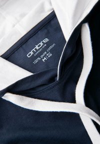 Námořnická modrá bavlněná mikina s bílým límcem a šňůrkami. Štítek označuje 100 % čistou bavlnu, velikost M, značka 'ombre'. Hladká textura.