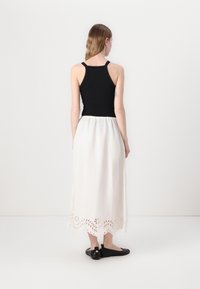 Schwarzes Tanktop mit dünnen Trägern, kombiniert mit einem fließenden weißen Rock mit gewelltem Saum und ausgeschnittenem Blumenmuster. Dazu werden schwarze Schuhe getragen.