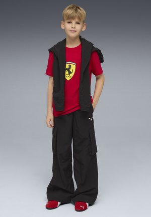 Niño joven de pie con una mano en el bolsillo, vistiendo una camiseta roja de Ferrari, pantalones cargo negros, chaqueta negra sobre los hombros y zapatos rojos de Puma.