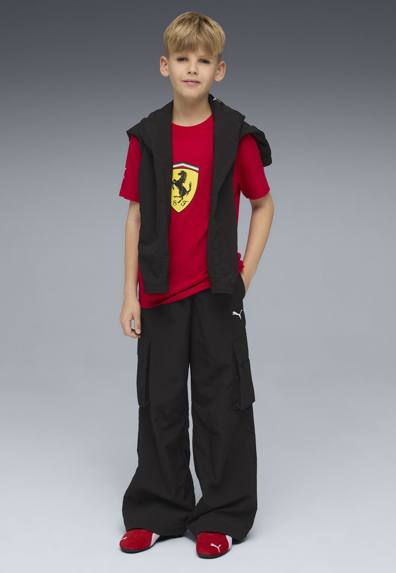 Junger Junge steht mit einer Hand in der Tasche, trägt ein rotes Ferrari-T-Shirt, schwarze Cargohosen, eine schwarze Jacke über den Schultern und rote Puma-Schuhe.