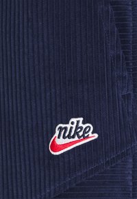 Nike-logopatch med röd swoosh och marinblå text broderad på mörkblå kordflorens.