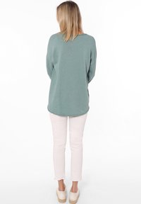 Hellgrüner Strickpullover mit entspannter Passform, kombiniert mit blassrosa Hosen, von hinten gezeigt. Legere Espadrilles runden das Outfit ab.