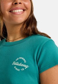 Teal geribbelde t-shirt met een ronde halslijn en korte mouwen, met een witte bedrukte logo waarop "The Original Billabong Since '73" staat.