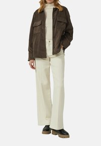 Vrouw in een bruine corduroy jas, crèmekleurige coltrui, crèmekleurige wijde broek en zwarte laarzen met dikke tan-zolen.