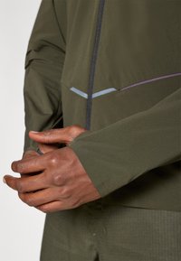 Ελαφρύ ελαιόκανο σακάκι με φερμουάρ μπροστά, που διαθέτει ανακλαστική ρίγα και υφασμάτινη υφή, που φαίνεται στα χέρια ενός ατόμου.