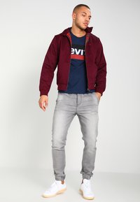 Chaqueta bomber burdeos con cremallera en la parte delantera, camiseta navy con logo rojo y blanco, jeans grises ajustados y zapatillas blancas con suela clara.