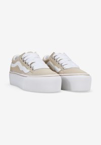 Zapatillas de lona beige con cordones blancos, una suela de goma blanca gruesa y un diseño ondulado en el lateral. Cuenta con ojales metálicos para los cordones.