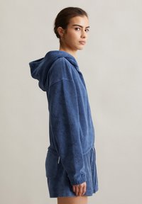 Felpa con cappuccio blu realizzata in morbido tessuto terry. Presenta un cordoncino in vita e tasche laterali. Vestibilità rilassata con maniche lunghe e superficie testurizzata.