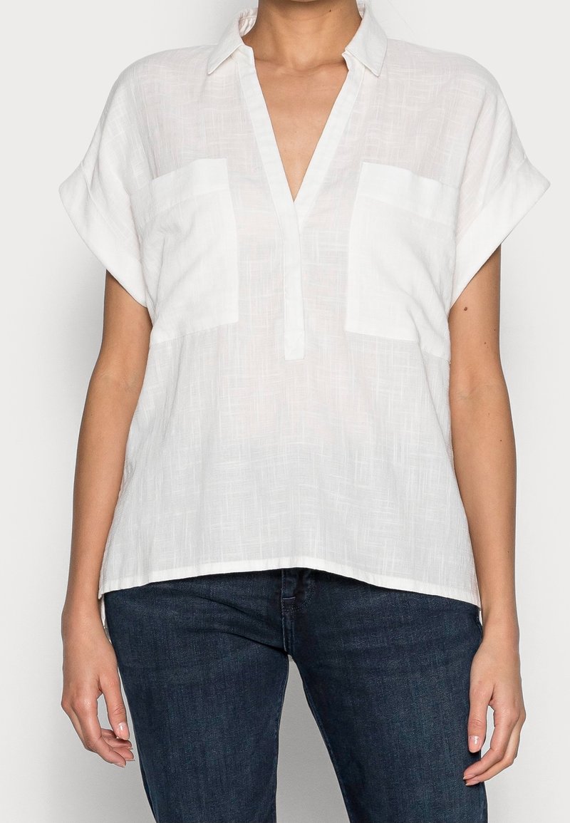Biała bluzka z krótkim rękawem, wykonana z lekkiego materiału, z dekoltem w serek i dwoma kieszeniami z przodu, o luźnym kroju i fakturowanym wzorze.