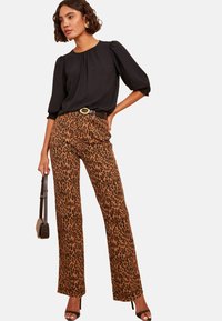 Donna con una blusa nera a maniche a sbuffo, pantaloni con stampa leopardata, cintura nera con fibbia dorata, tacchi neri, in possesso di una piccola borsa marrone.