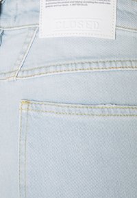 Jean en denim bleu clair en gros plan montrant la poche arrière, les coutures jaunes et une étiquette en tissu blanche avec le nom de la marque embossé "CLOSED."