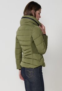 Chaqueta acolchada de color verde oliva con un alto cuello, diseño acolchado y paneles de punto laterales para mayor textura, combinada con jeans de mezclilla oscura.