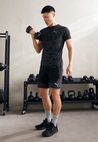 Maglietta sportiva nera con motivo, pantaloni corti neri con dettagli in rete, calze corte grigie e scarpe da allenamento nere; sollevando pesi in una palestra.