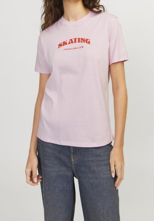 Kobieta ubrana w jasnoróżowy t-shirt z czerwonym napisem "SKATING UNAVAILABLE ATM" i niebieskie dżinsy, stoi na tle jednolitego jasnego tła.